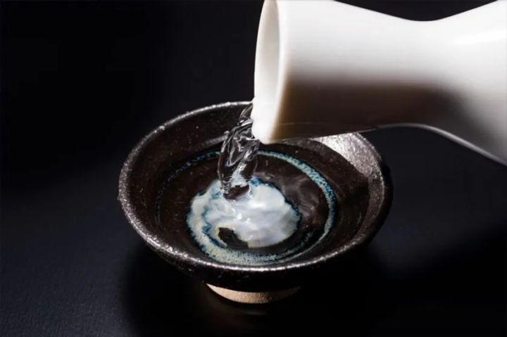白い徳利から黒釉の盃に日本酒を注ぐ瞬間をクローズアップした暗背景のシズル写真