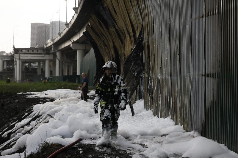 A firefighter covered in foam walks past a burned fence underneath an overpass.