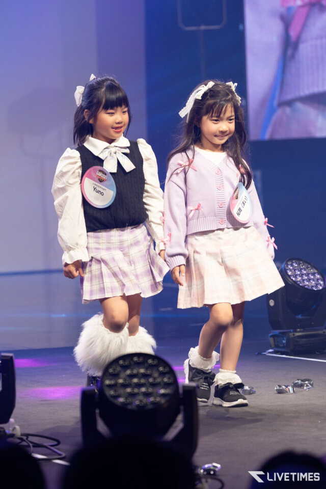 Two young girls on a stage wearing pastel outfits and badges, smiling during a performance.