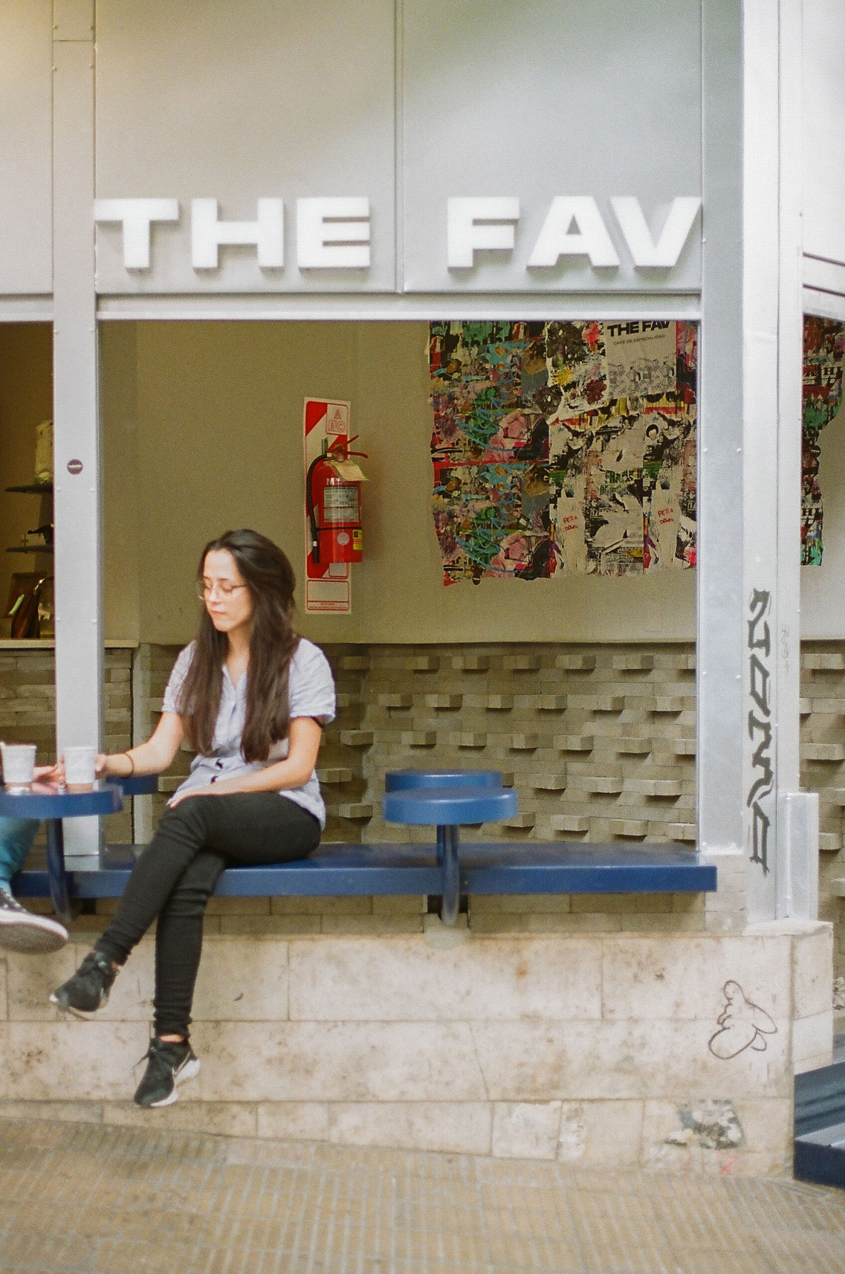 Woman with long dark hair sits on a blue bench outside a storefront labeled THE FAV, with a red fire extinguisher and colorful posters on the wall behind her.