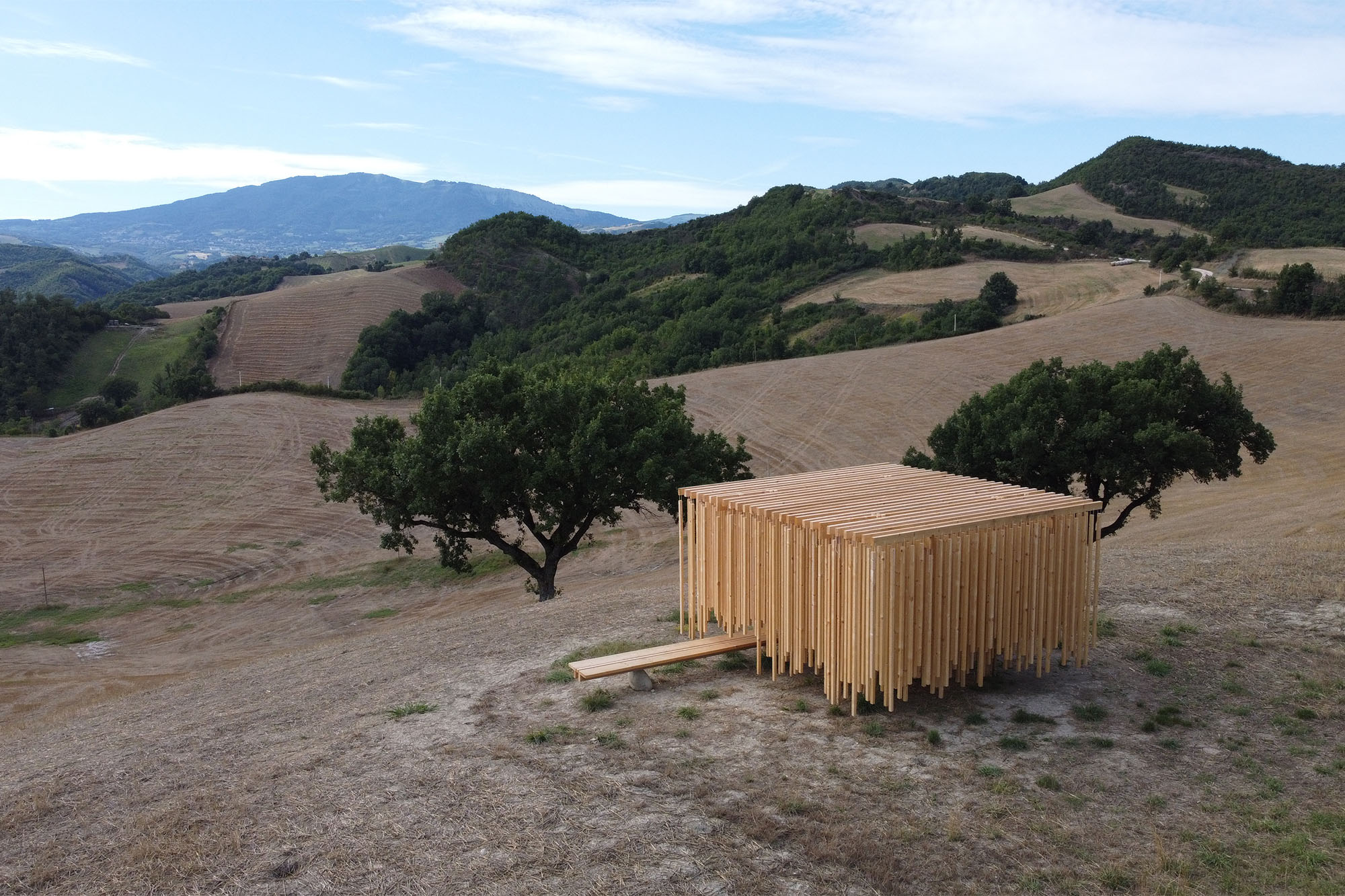 Wide hillside landscape with dry, harvested fields, green trees, and a wooden slat-framed structure in the foreground