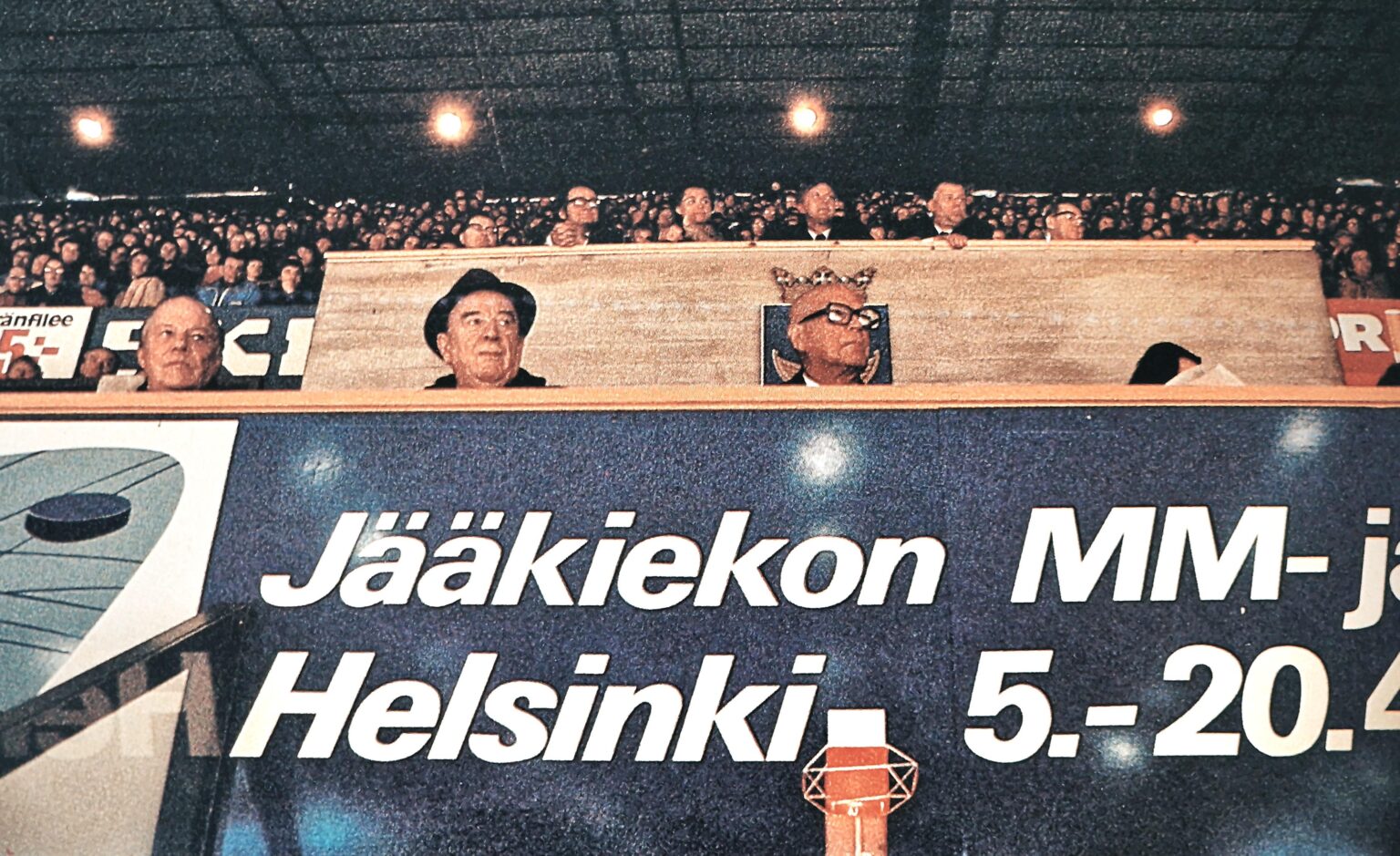 1974年、ヘルシンキでアイスホッケーの世界選手権を観戦するフィンランドの神ウルホ・カレヴァ・ケッコネン（ハゲ、でっかいメガネ）。写真家が正しい角度を選んだ。彼の頭の後ろにはヘルシンキの紋章と王冠がある。