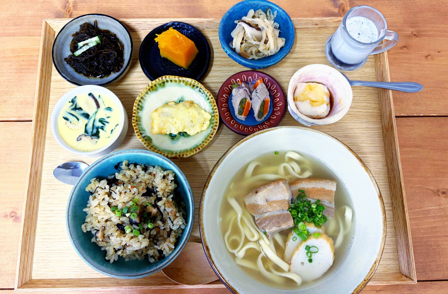<div class="caption">一番人気の「ほっこり定食」。小鉢は(左上から時計回りに)モズク酢、カボチャ煮、千切りイリチー、黒ごまプリン、ジーマーミ豆腐、インゲンの肉巻き、バサの天ぷら、茶碗蒸しの8品が並ぶ。小鉢のメニューは定期的に変わる</div>