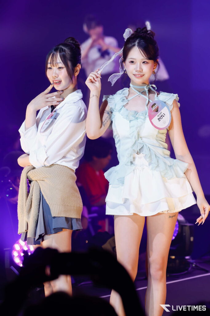 Two young women posing on a purple-lit stage, smiling at the audience with badges on their outfits.