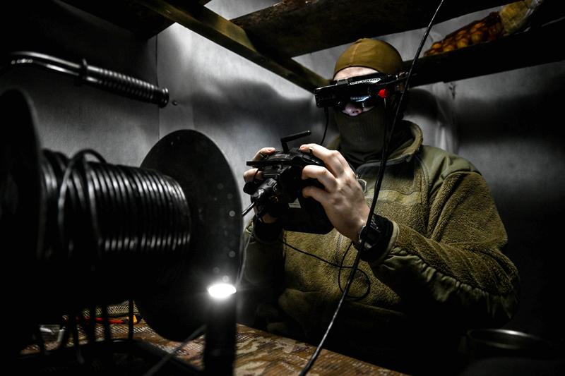 A man in khaki and a headset holds a controller in a small grey enclosed space with a black spool of cable in front of him