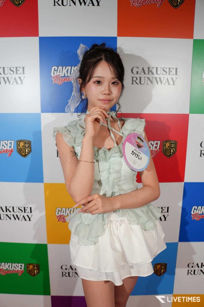 Young woman posing at a Gakusei Runway event, wearing a pale green ruffled top and white skirt, against a colorful checkered backdrop.