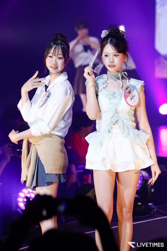 Two young women perform on stage under purple lighting, posing for the audience at a concert.