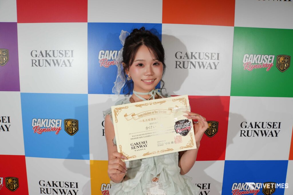 Smiling young woman in a light dress holding a certificate at a colorful GAKUSEI RUNWAY backdrop.