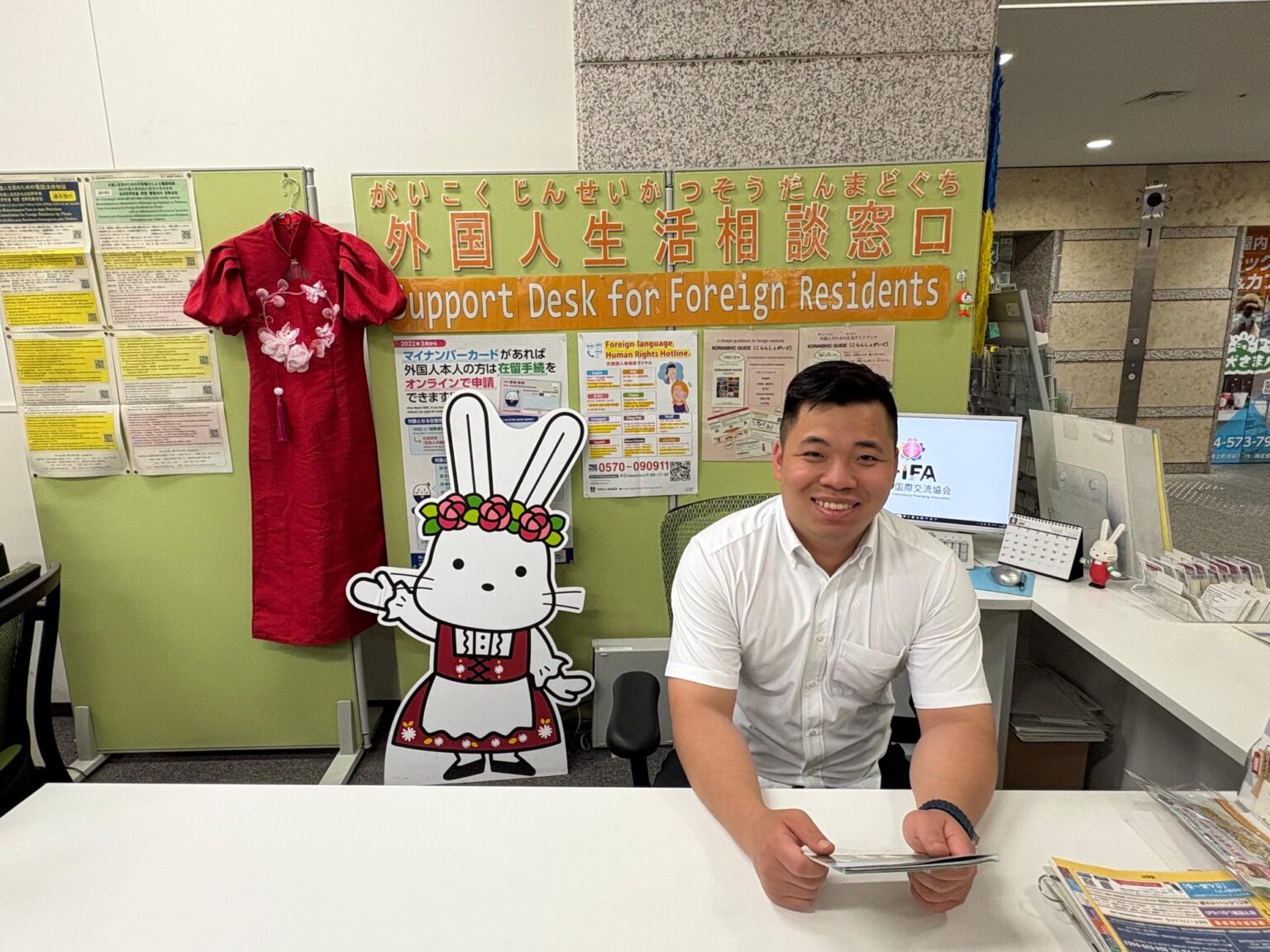 外国人生活相談窓口(Support Desk for Foreign Residents)/福島市公式ホームページ 外国人生活相談窓口でクインさんが笑顔で座っている写真。ベトナムの民族衣装の赤いアオザイとももりんのパネルが飾られている。