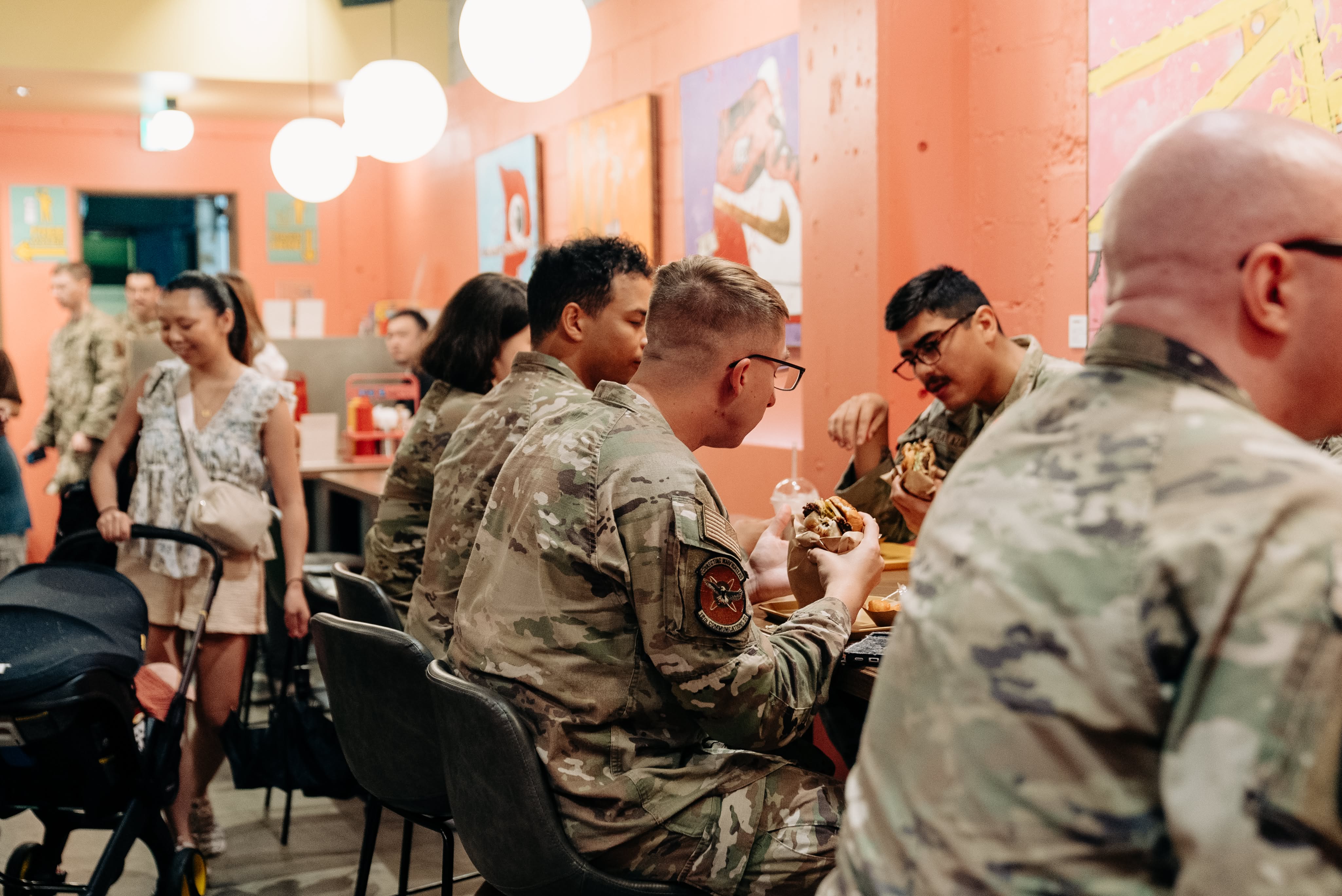 People in military uniforms eating burgers at a casual restaurant with orange walls and modern art.