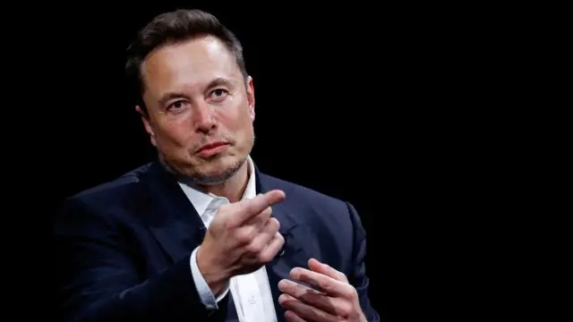 ダークスーツ姿のマスク氏が前方を指差している