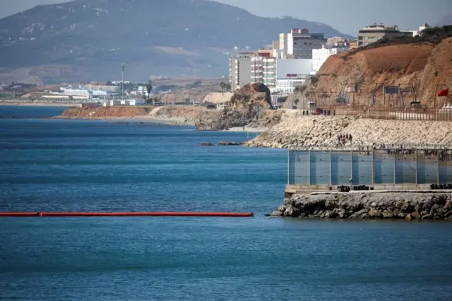 海に突き出た岩に国境フェンスが立ち、その先の洋上に赤いまっすぐなブイが浮いている