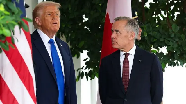 トランプ氏とカーニー氏が並んで立っている。トランプ氏は遠くの方を見ており、カーニー氏はトランプ氏の顔を凝視している。周囲にはアメリカ国旗とカナダ国旗が並べらている